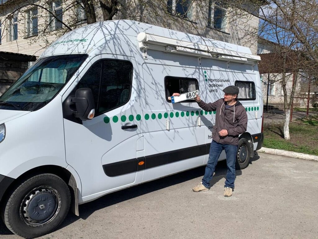 Man in a gray jacket and cap extends a card toward a white service van with green dots parked on a street.