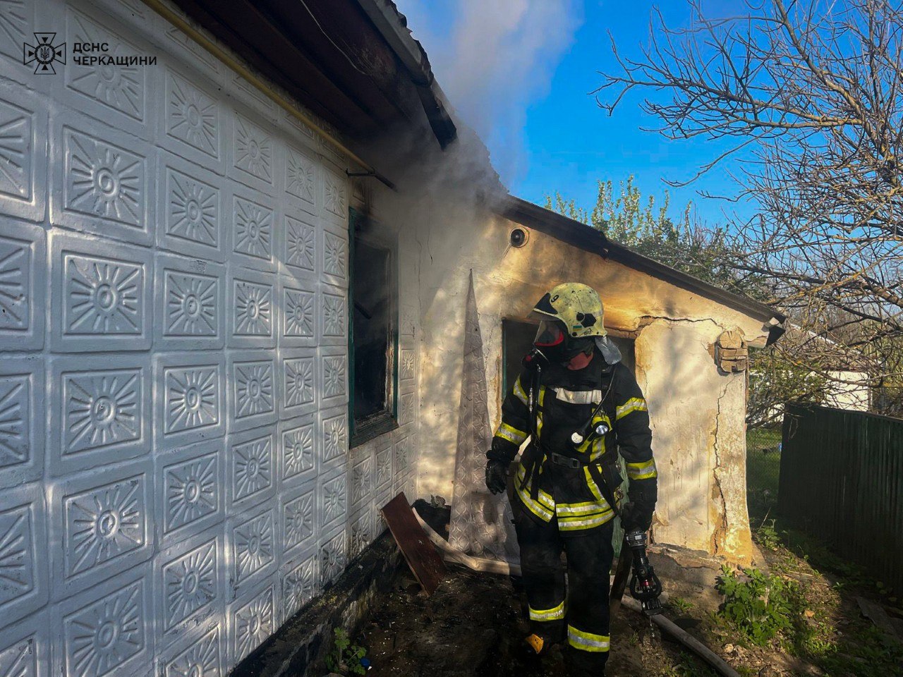 Firefighter in protective gear exits a burning, smoke-filled house with charred exterior walls and a window open to the left.