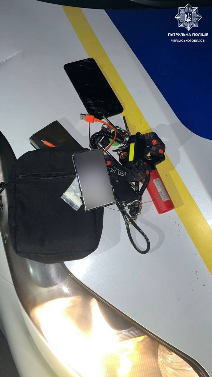 Gear laid on the hood of a police car: a black bag, a smartphone, charging cables, a red flashlight, and other small equipment; police emblem visible in the top-right corner.
