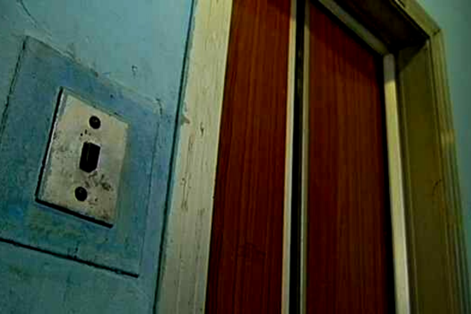 Worn metal light switch plate on a blue wall beside a brown wooden doorway trim, looking old and scratched.