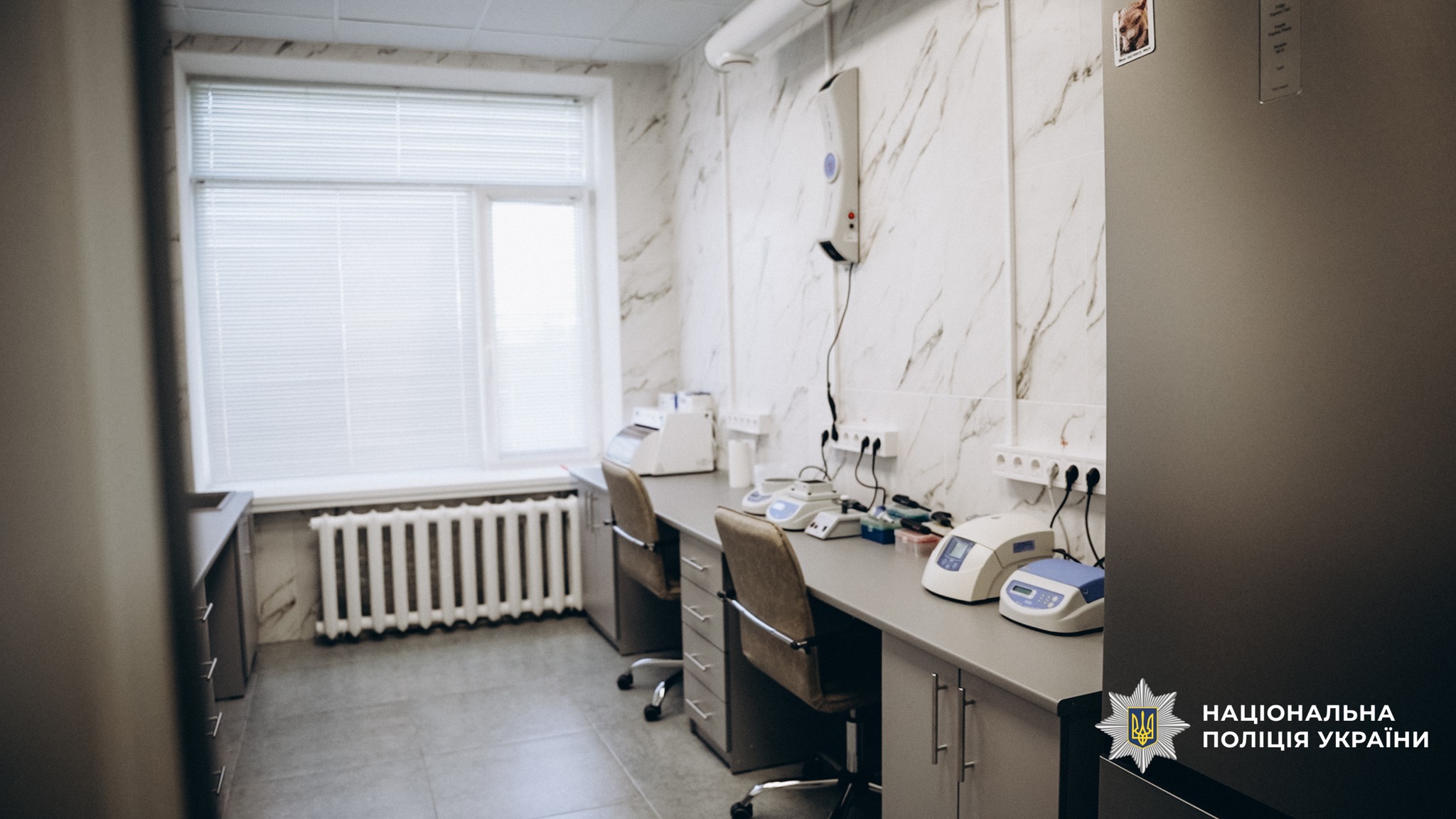 Laboratory or clinical workspace with medical devices on a long counter under marble-wall panels and a large window with blinds on the left, radiator beneath the window, and chairs at the counter.