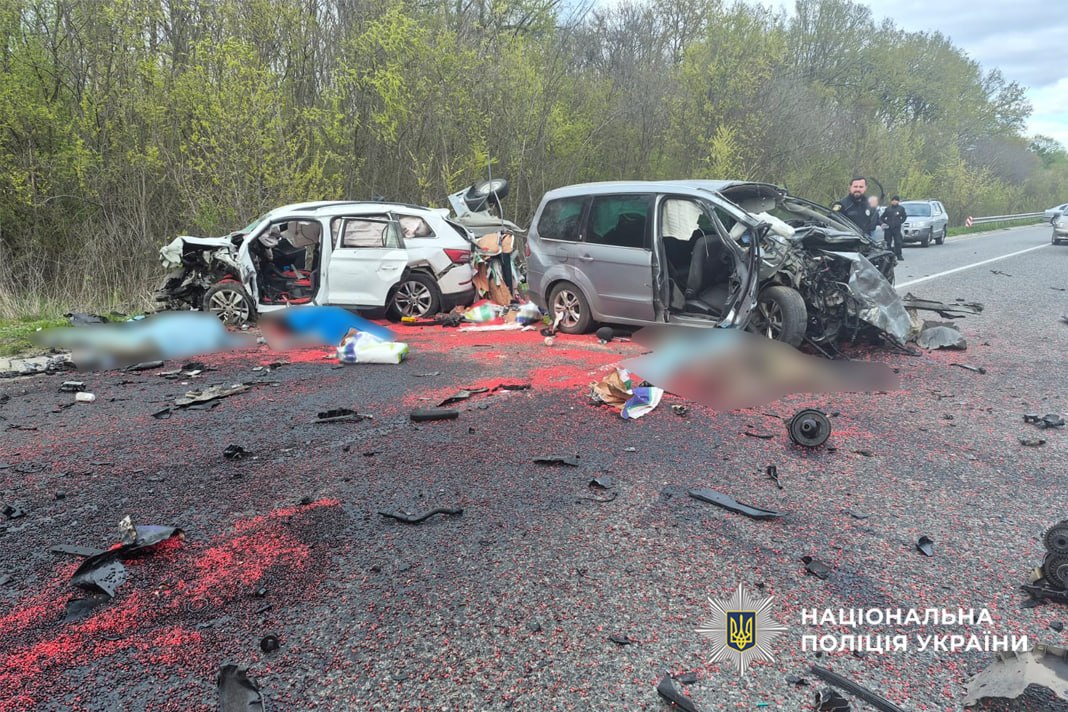 Multivehicle crash on a highway with two heavily damaged vehicles, debris and red fluid on the road, emergency responders nearby; Ukrainian police logo visible.