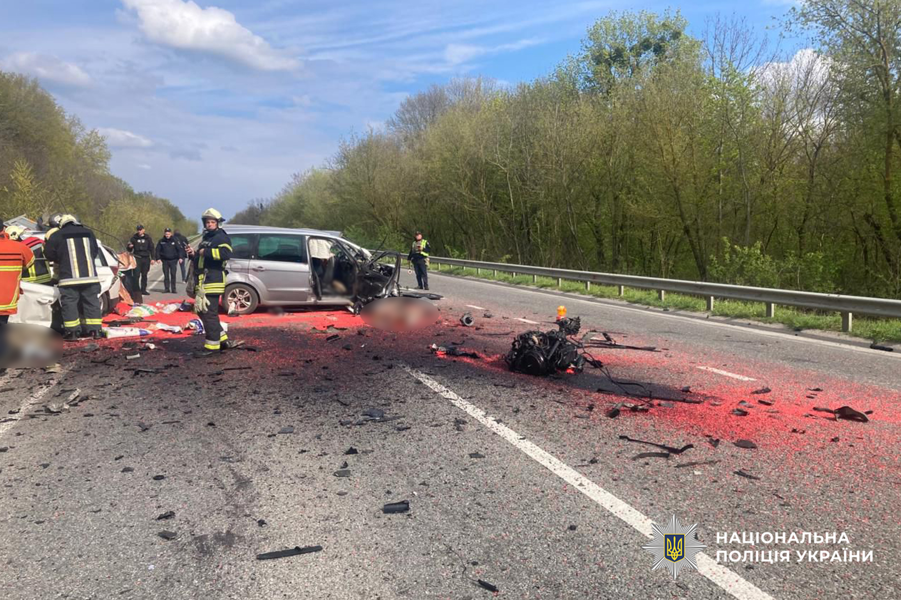 Emergency responders at a highway crash scene with a severely damaged van, scattered debris, and a red-stained road surface.