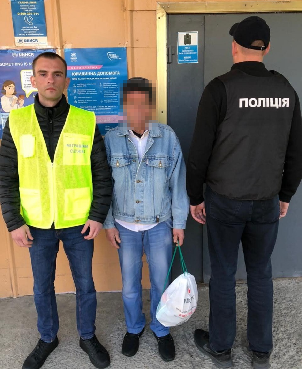 Two uniformed officers stand with a man whose face is blurred outside a building with blue posters; the center person holds a white plastic bag.