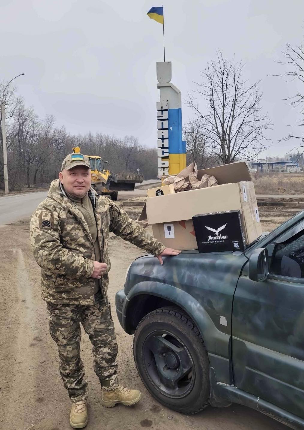 Soldier in camouflage uniform stands beside a pickup truck with boxes loaded on the hood; Ukrainian flag visible in the background.