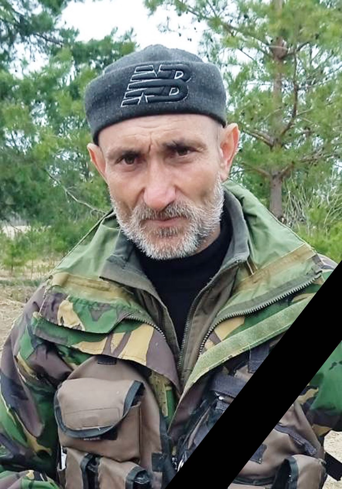 Man in camouflage jacket outdoors with a black diagonal mourning ribbon across the photo.