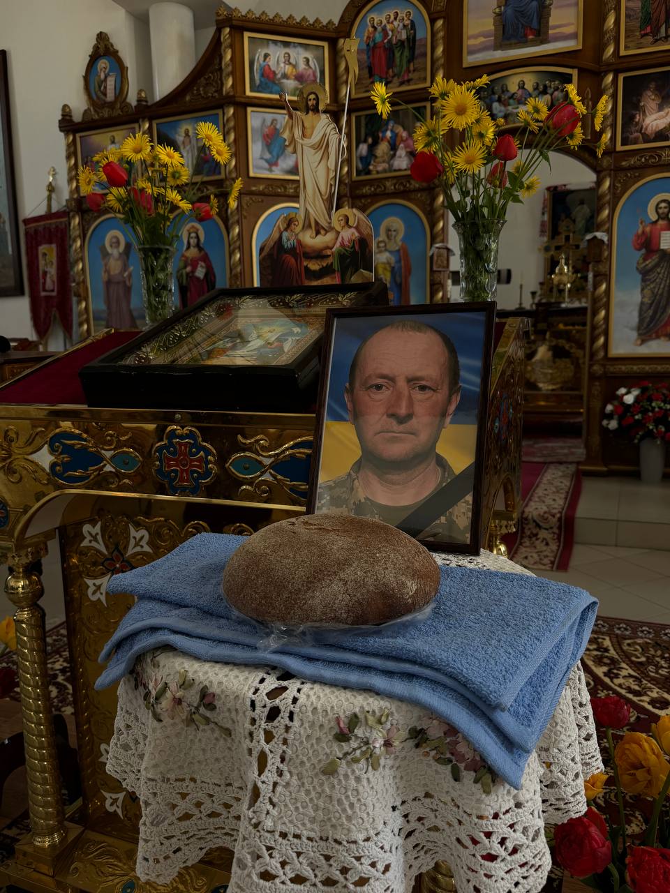 Memorial display in a church: framed photo of a man in military uniform on a lace-covered table with a loaf of bread, blue cloth, and yellow flowers; ornate icons in the background.
