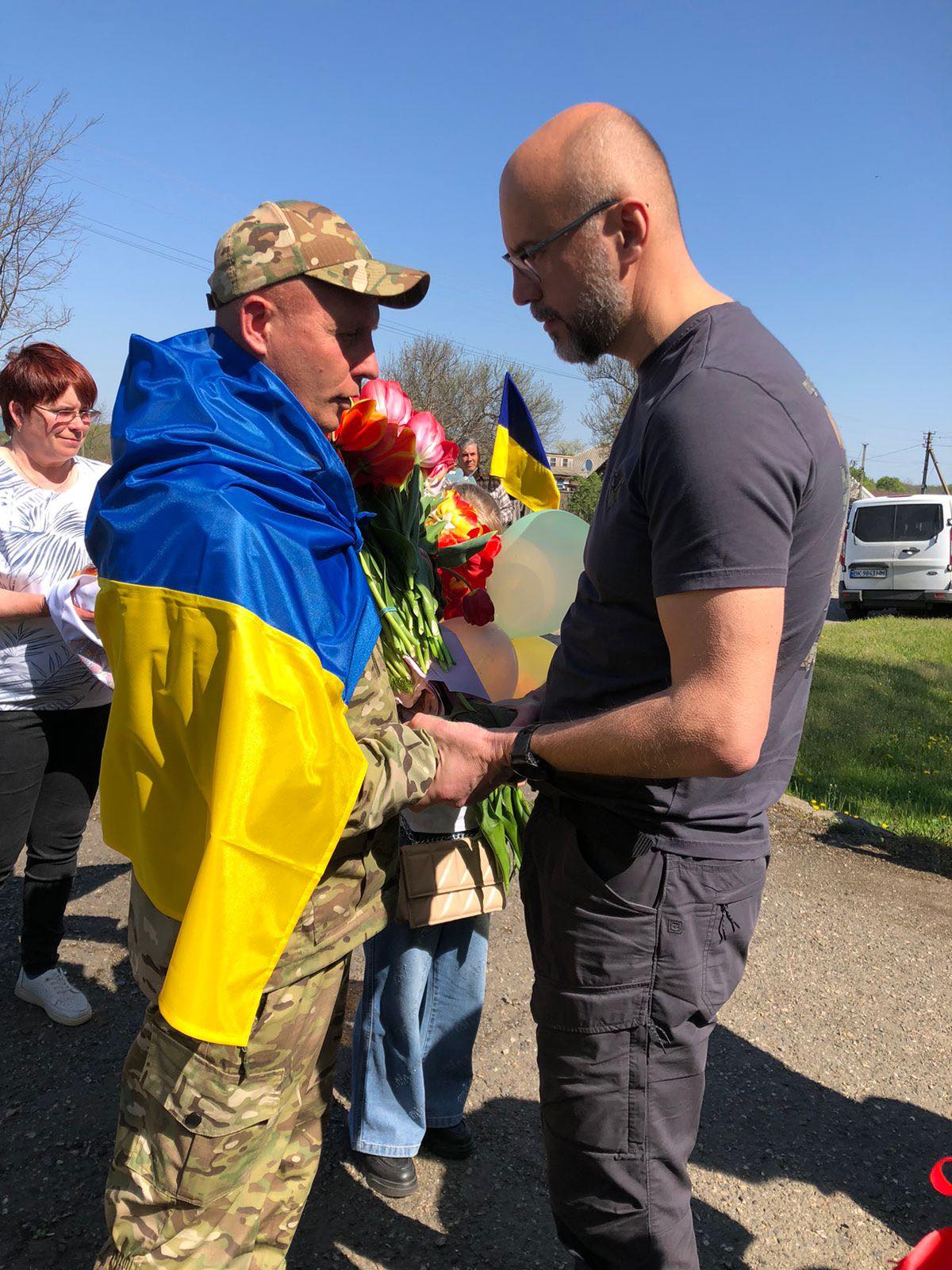 Soldier in camouflage with a blue-yellow Ukrainian flag draped over his shoulders accepts a bouquet while a man in a dark shirt shakes his hand outdoors, onlookers and a Ukrainian flag in the background.