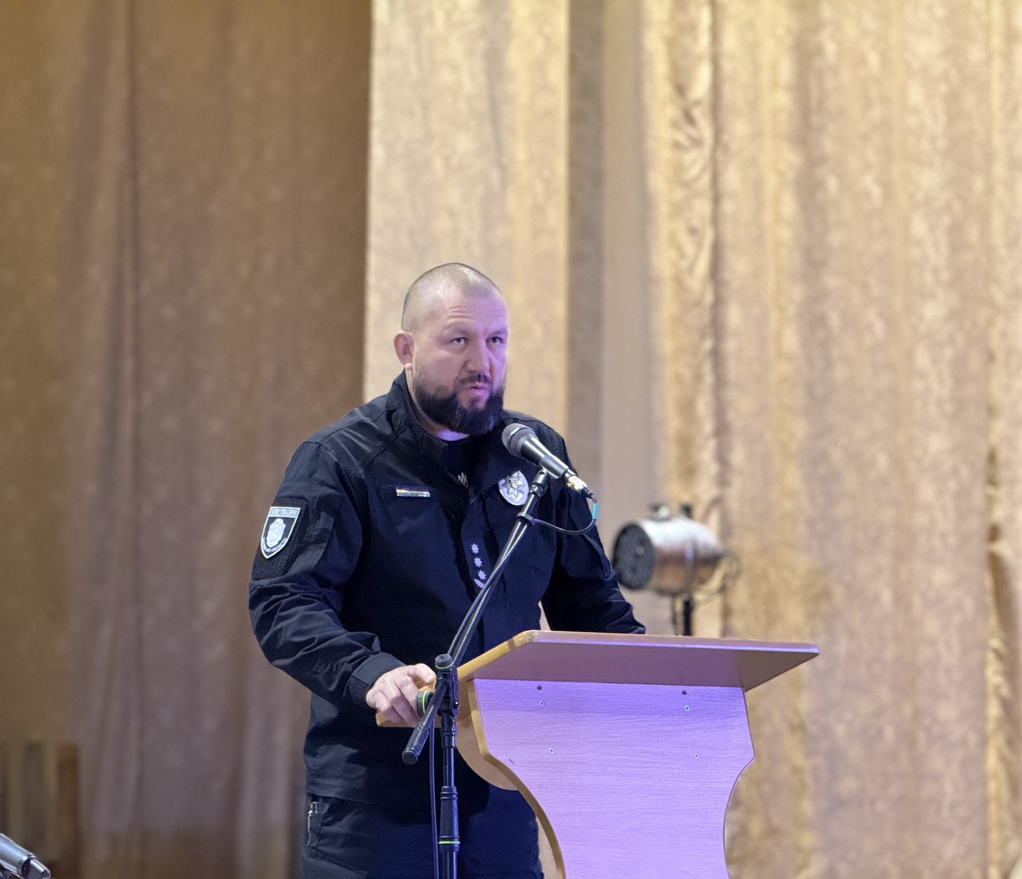 Bearded man in a police uniform delivering a speech at a wooden podium with a microphone, in front of curtain backdrop.