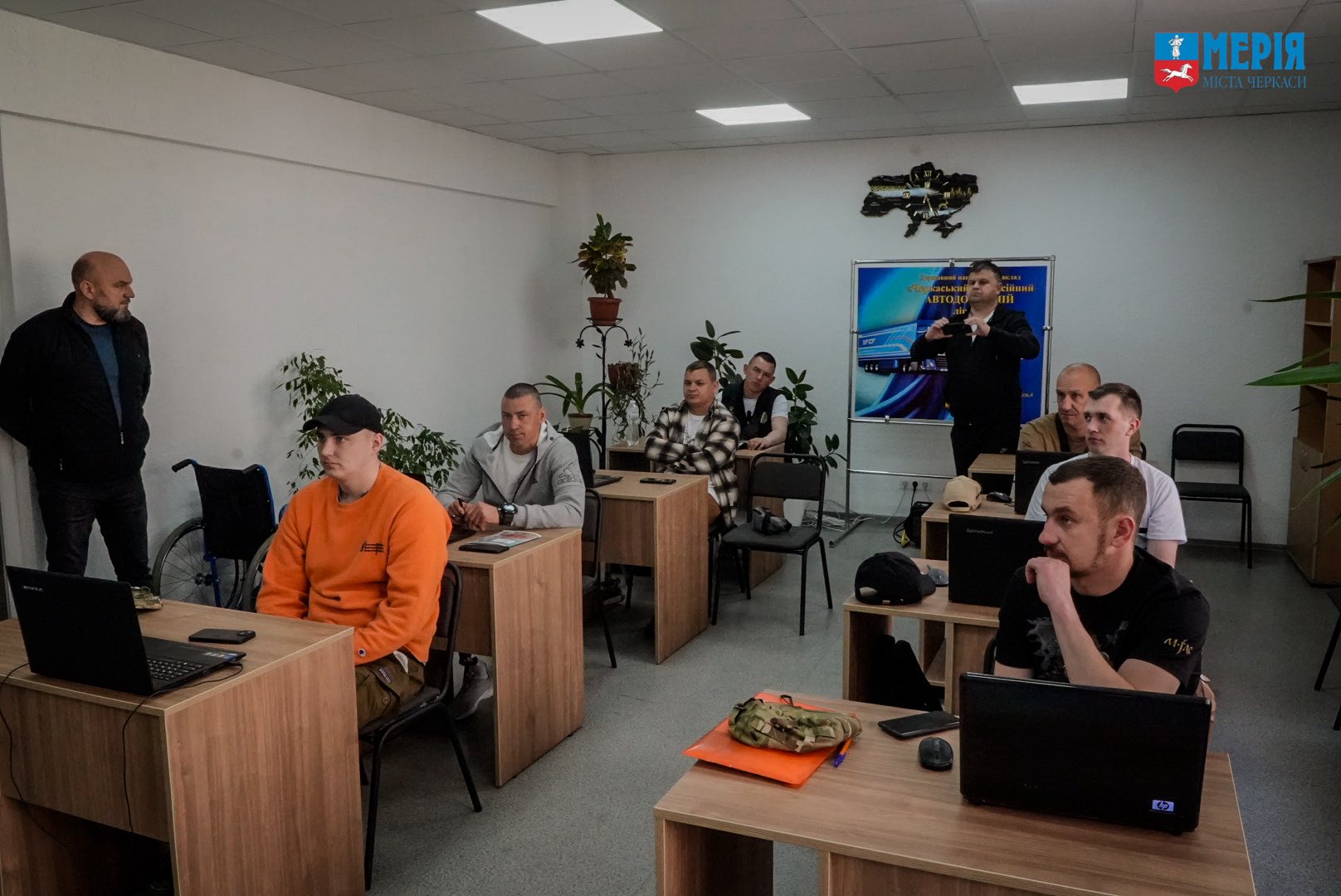 Instructor gives a presentation to a classroom of men at wooden desks with laptops; plants line the back wall and a poster behind the speaker.