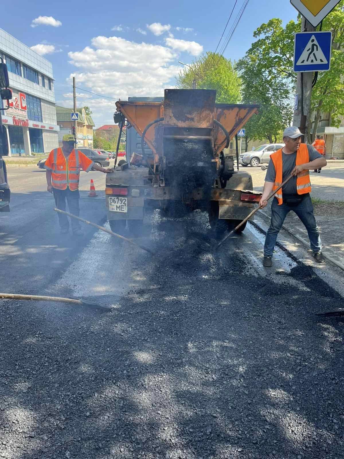 Road workers in orange vests spreading and leveling fresh asphalt behind a dump truck on a city street under a clear sky.