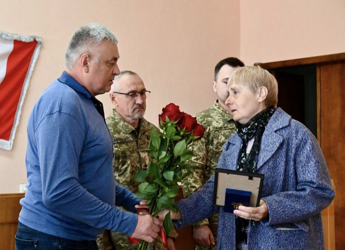 Man in a blue sweater hands red roses to a woman in a blue coat during a ceremony, with soldiers in camouflage in the background.