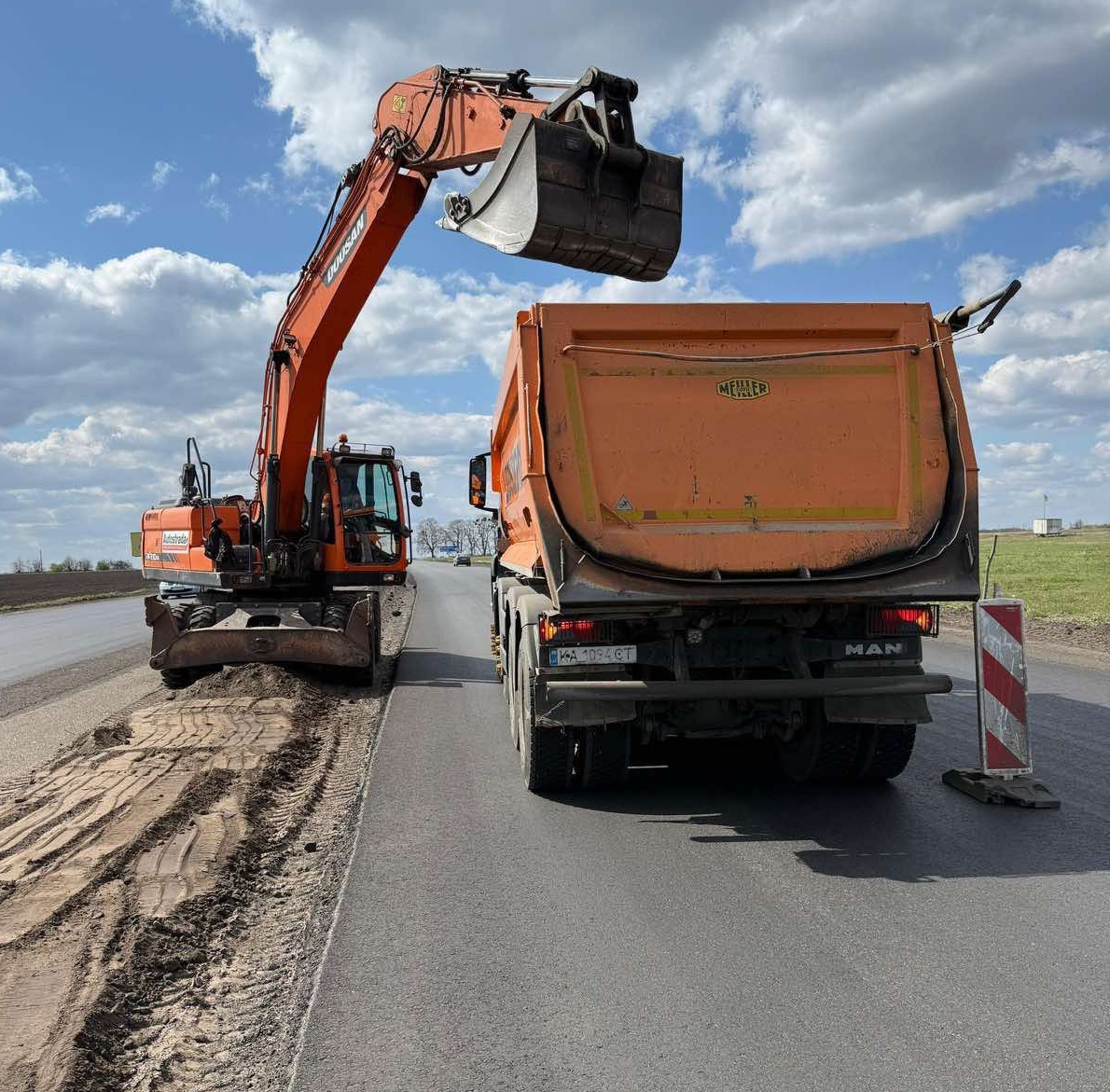 Orange excavator with raised bucket transfers soil into a large orange dump truck on a rural road at a roadwork site under a cloudy sky.