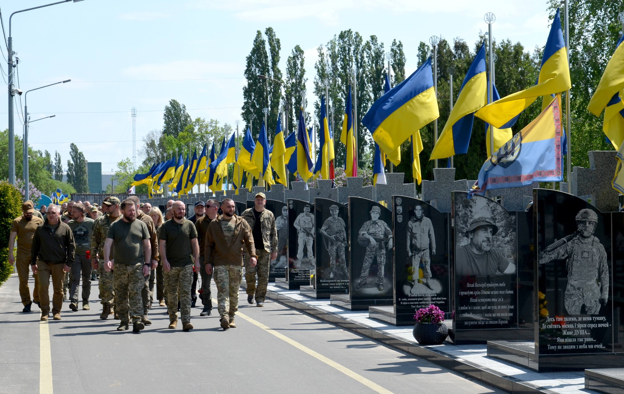 Soldiers in uniform marching along a road beside a row of blue-and-yellow Ukrainian flags atop flagpoles and a line of memorial panels with photos and inscriptions.