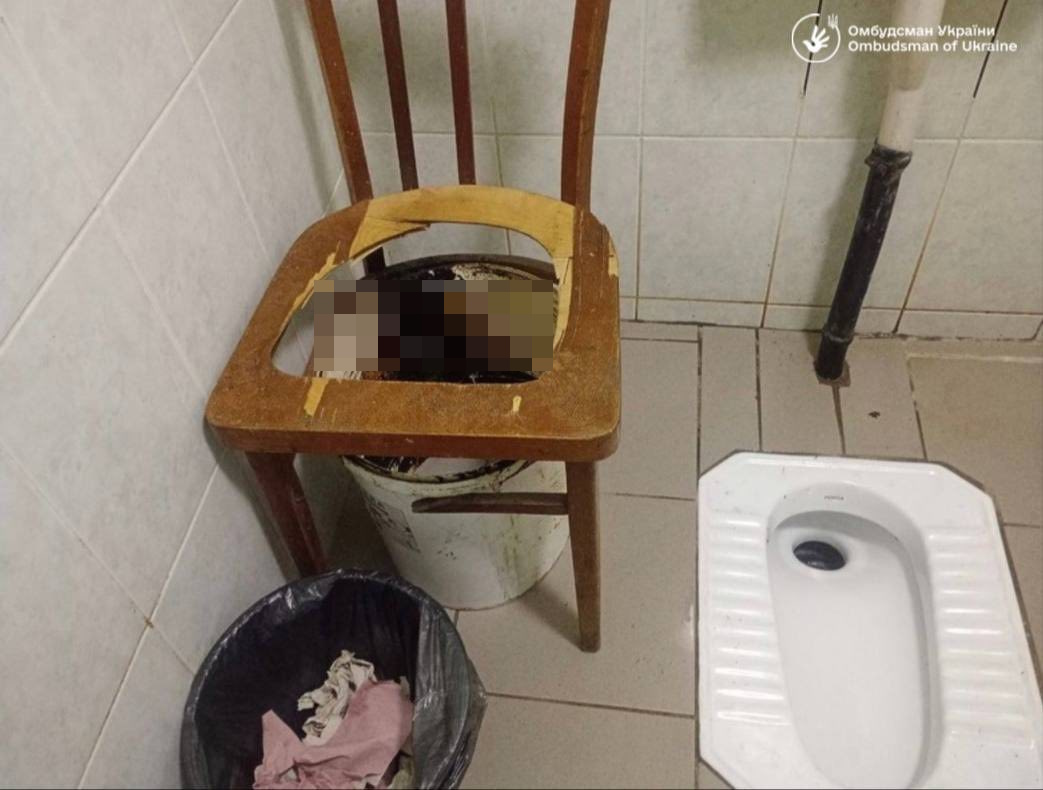 Dilapidated bathroom with a wooden chair placed over a bucket, a white squat toilet on the floor, and a trash can with rag scraps nearby.