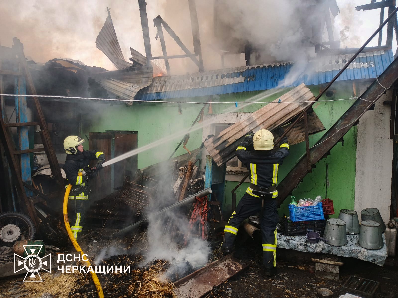 Firefighters in protective gear spraying water at a burning, collapsed building with smoke and charred debris.
