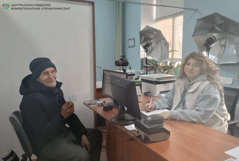 Elderly man in a black coat and hat sits across from a young woman at a desk with a computer in a studio-like office with lighting equipment behind them.