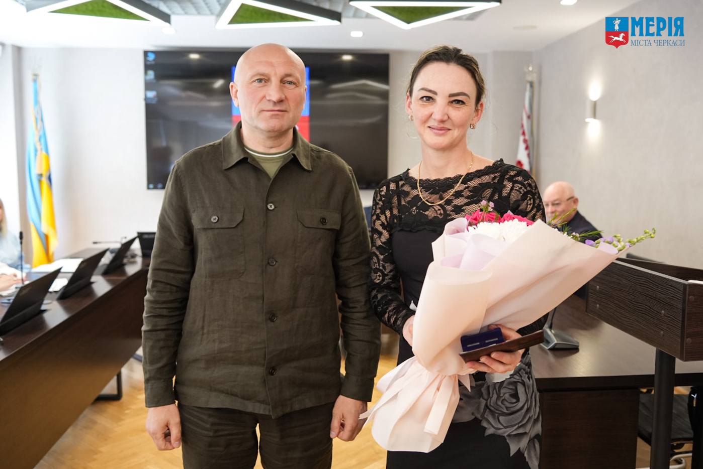 Man in a green utility-style jacket stands next to a smiling woman in a black lace dress, who holds a large bouquet of pink flowers after a ceremony.