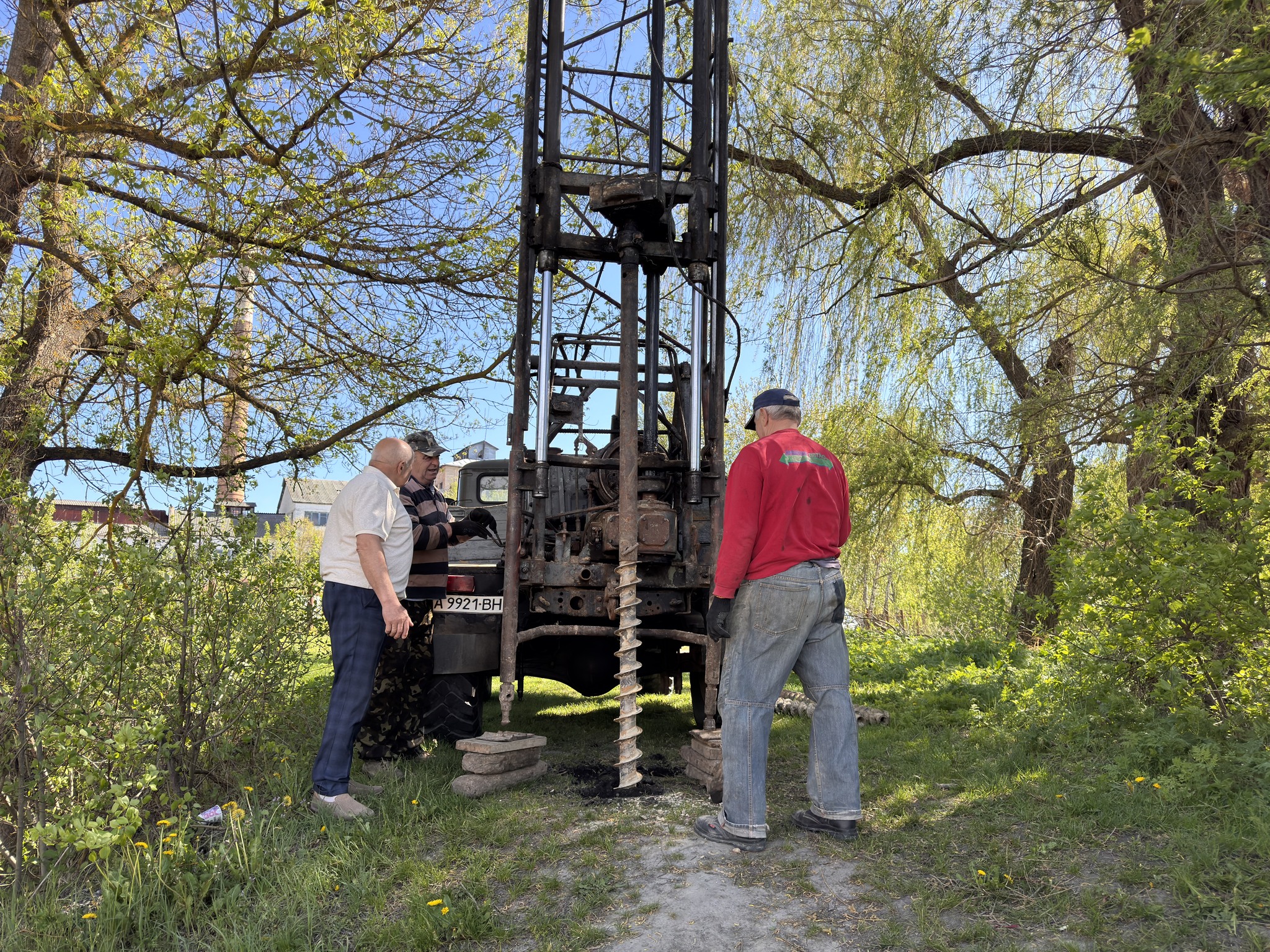 Three men inspect a rusted drilling rig outdoors among trees on a sunny day.