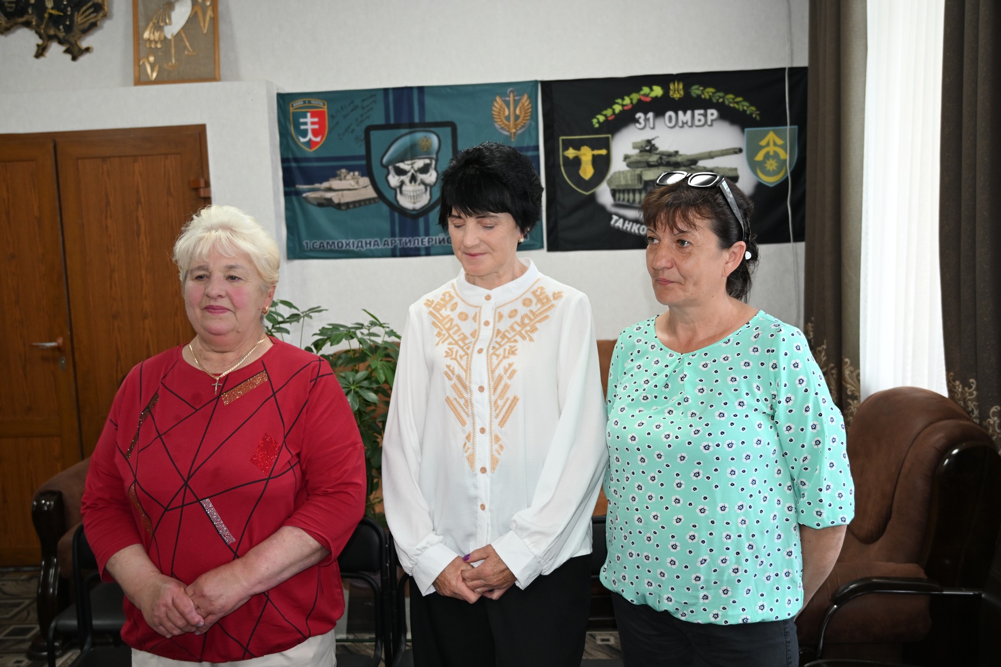 Three women stand indoors, left in a red patterned top, center in a white embroidered blouse, and right in a turquoise pattern blouse with glasses on her head, posing for a photo in front of military-themed banners.