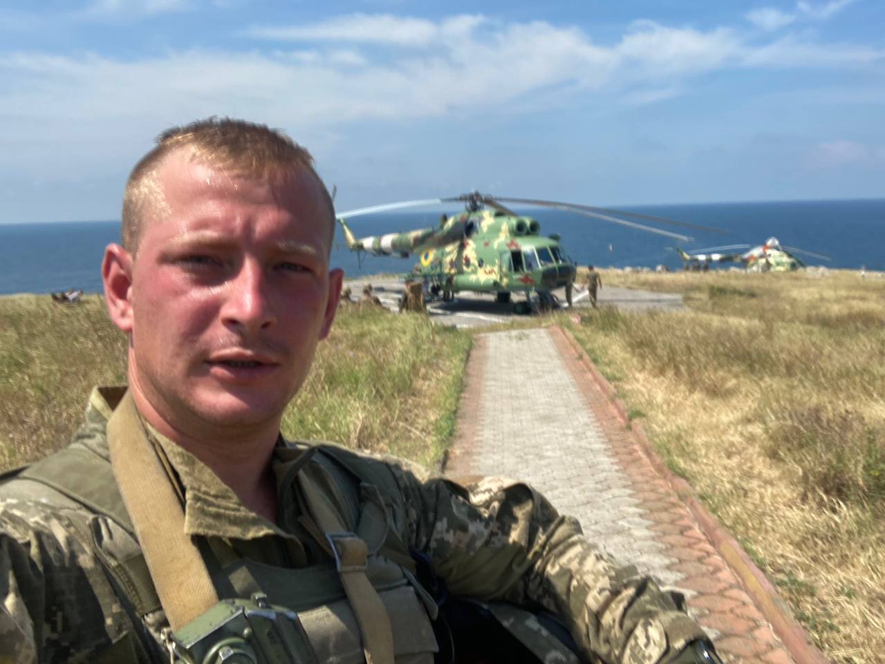 Soldier in camouflage taking a selfie on a coastal walkway with a green helicopter parked nearby by the shore.