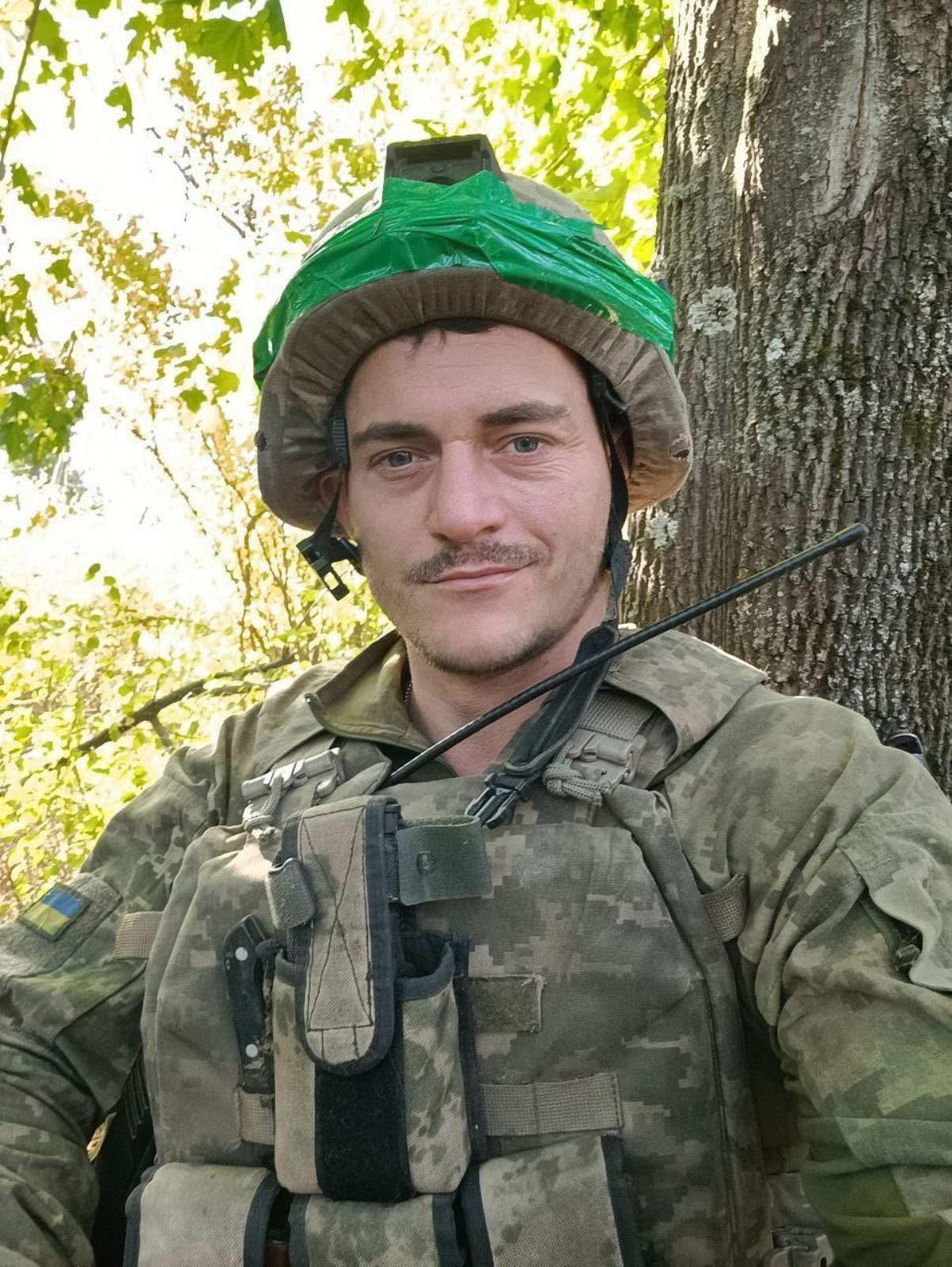 Military personnel in camouflage gear posing in a forest, wearing a helmet with a green plastic cover and tactical vest with gear.