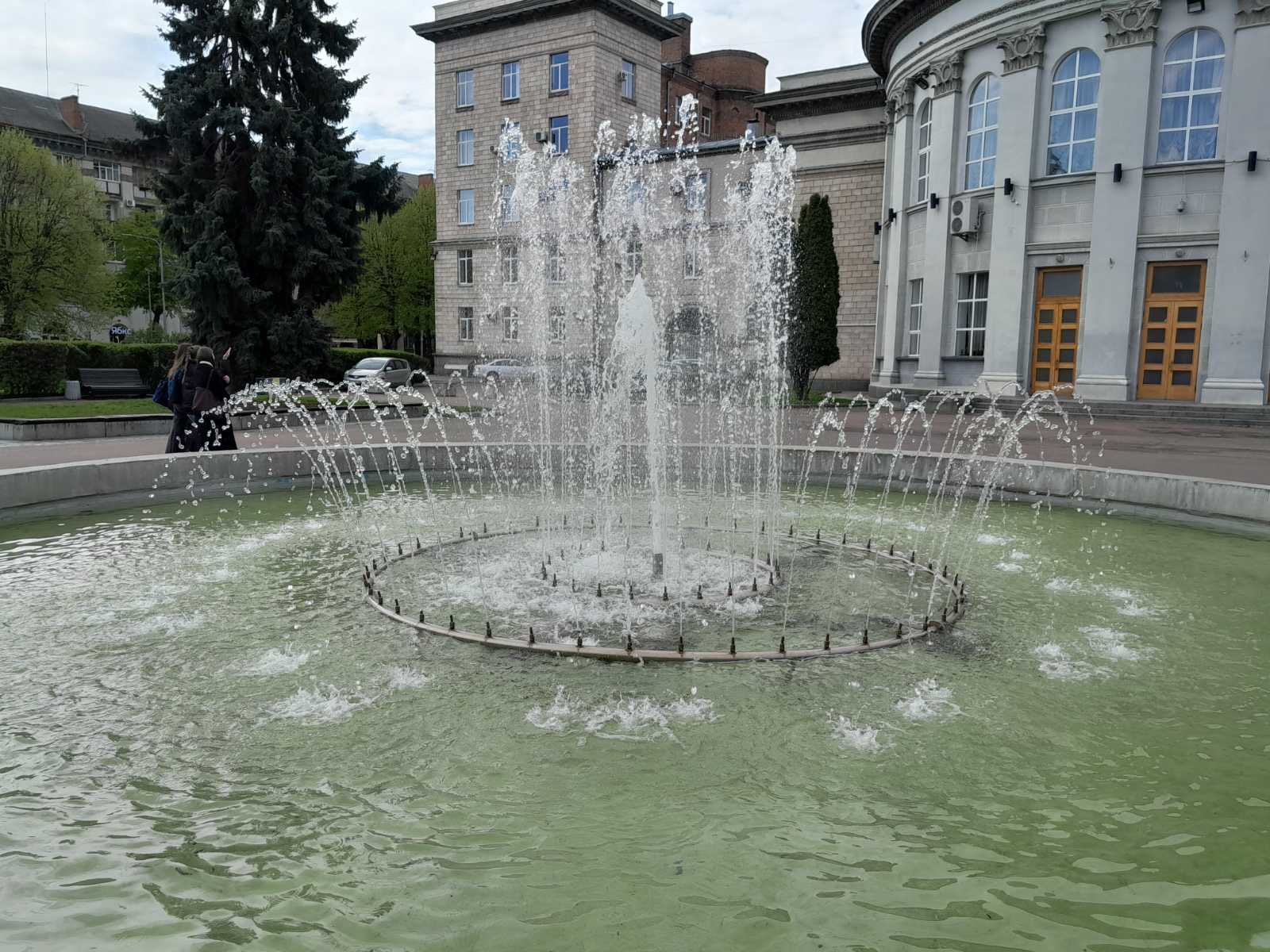 Fountain with circular jet sprays in a courtyard, with a stately building and trees in the background; a group of people walk nearby in the scene.