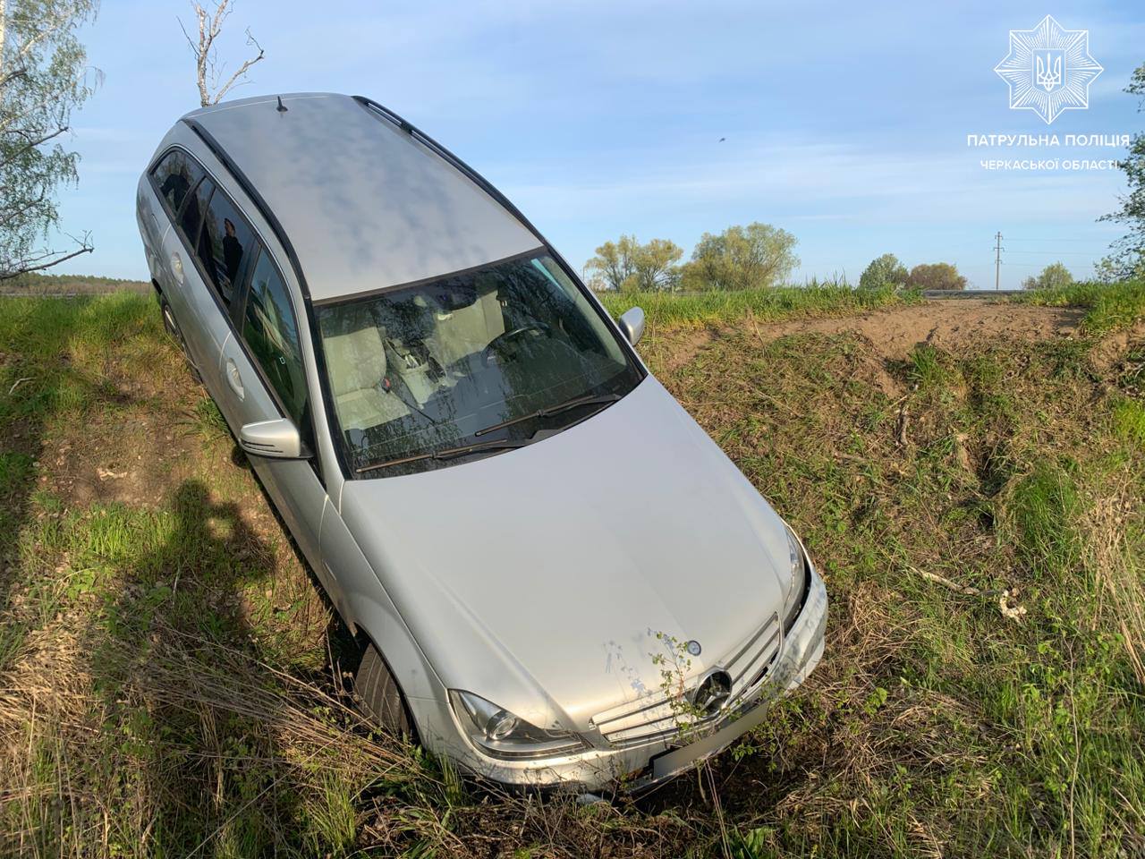 Silver sedanendra car stuck in a grassy ditch beside a rural road, seen from above; police logo in top-right corner.