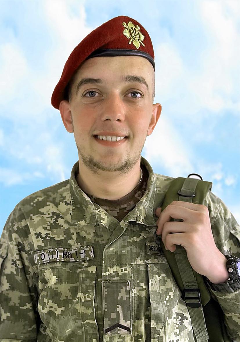 Smiling soldier in camouflage uniform wearing a red beret and carrying a green backpack against a blue sky background.