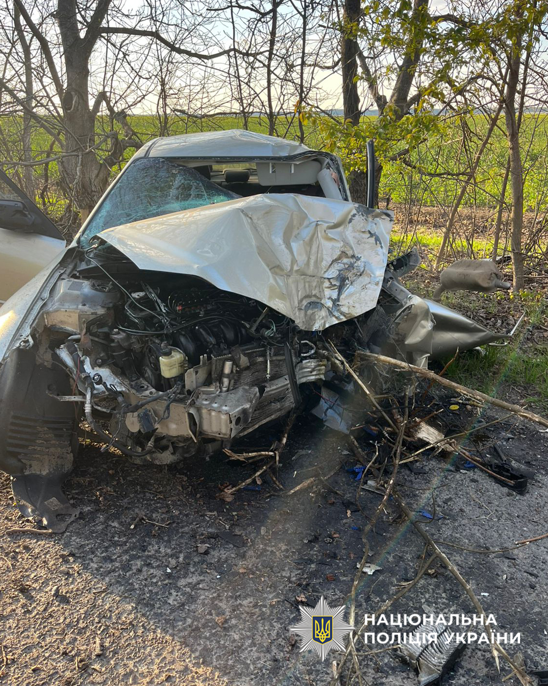 Severely damaged silver sedan with crumpled hood and shattered windshield after a crash, debris around on a roadside, police watermark bottom right.