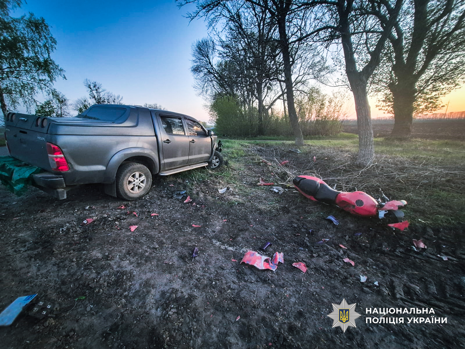 Silver pickup crashed in a rural field with a red motorcycle lying on its side and debris scattered nearby as dusk falls.