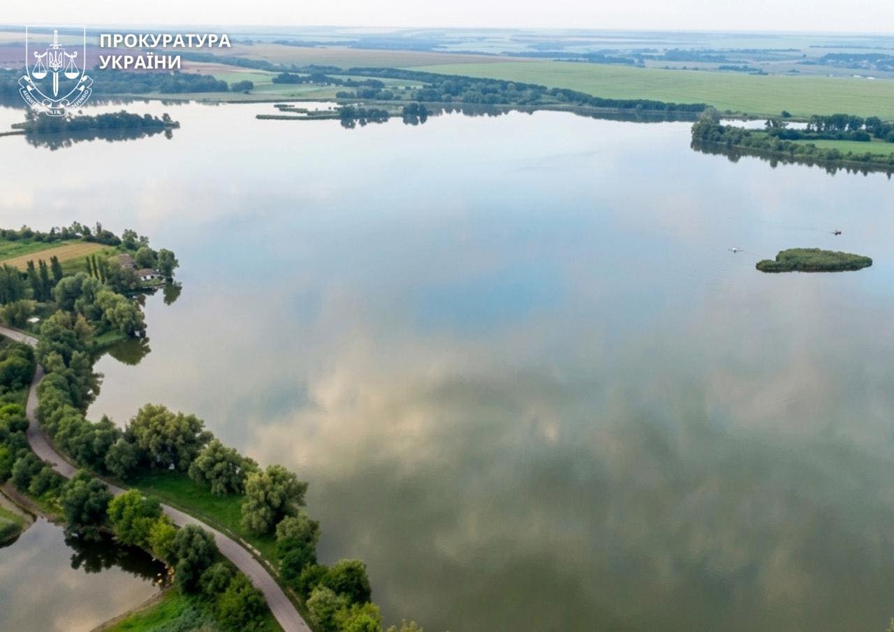 Aerial view of a wide, calm river with green fields and a tree-lined path along the left shore; Ukrainian Prosecutor's Office watermark in the top-left corner.