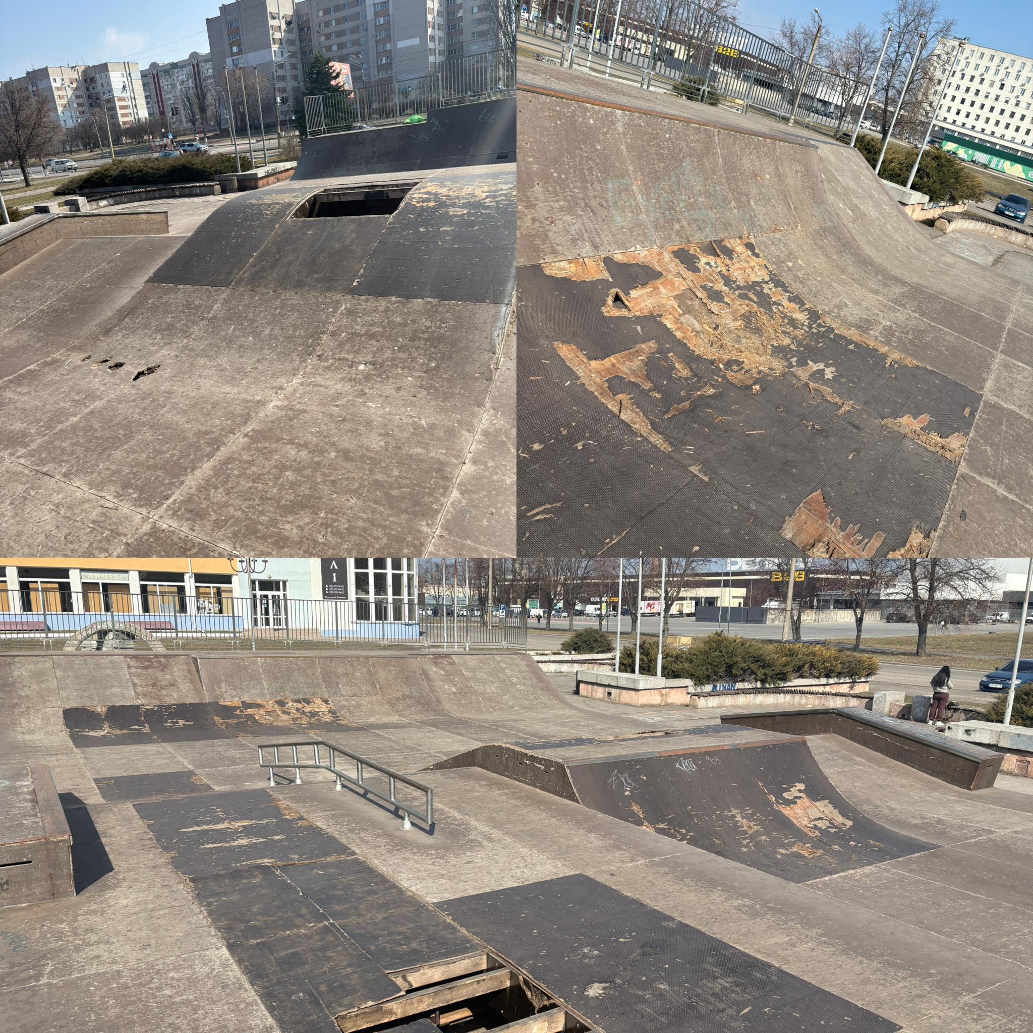 A vacant outdoor skatepark with cracked concrete ramps and a large hole in the foreground, surrounded by metal railings and urban buildings in the background.
