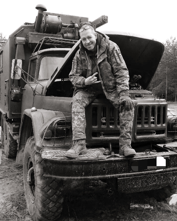 Man in camouflage sits on the hood of a large rugged truck with its hood open, posing for the camera.