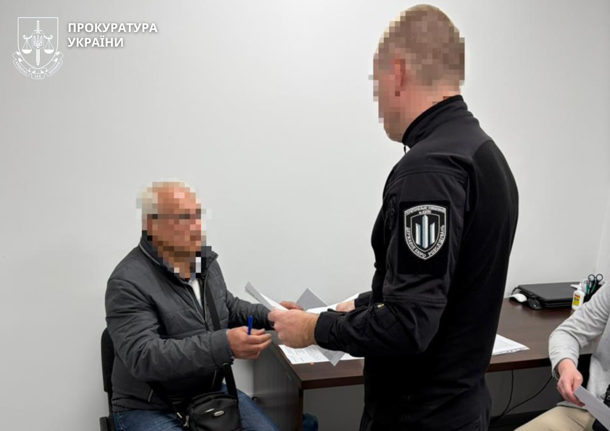 Two men exchange documents in an office; one seated, one standing, faces blurred; Ukrainian prosecutor’s emblem in the top-left corner.