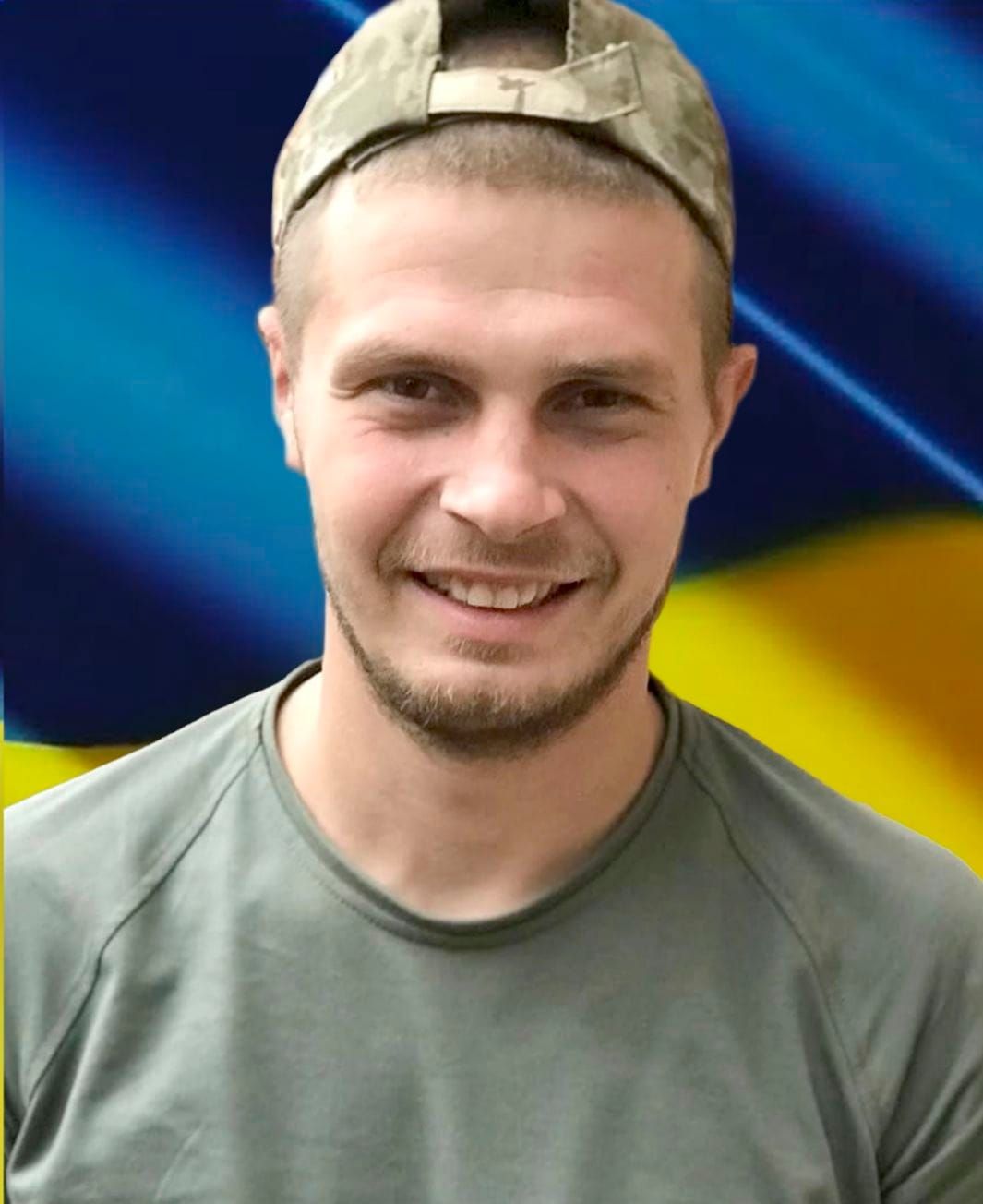 Smiling man wearing a backward camouflage cap and olive-green shirt, with a blue and yellow abstract background.