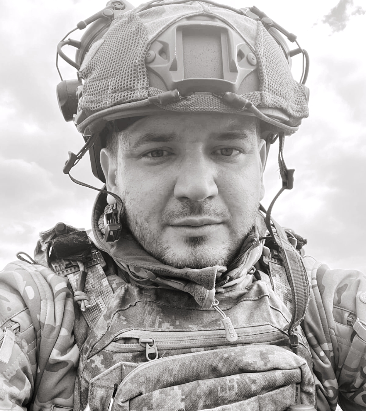 Close-up portrait of a soldier wearing a tactical helmet, headset, and camouflage vest, looking at the camera.