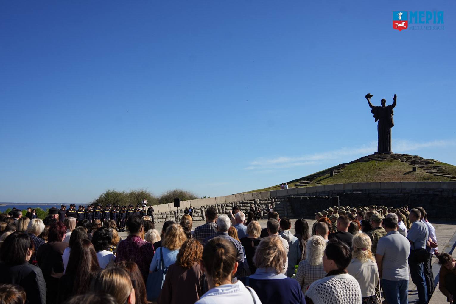 Crowd gathered at a memorial site in front of a hilltop statue under a clear blue sky, with the statue raising its arms in triumph and a lined group at left.
