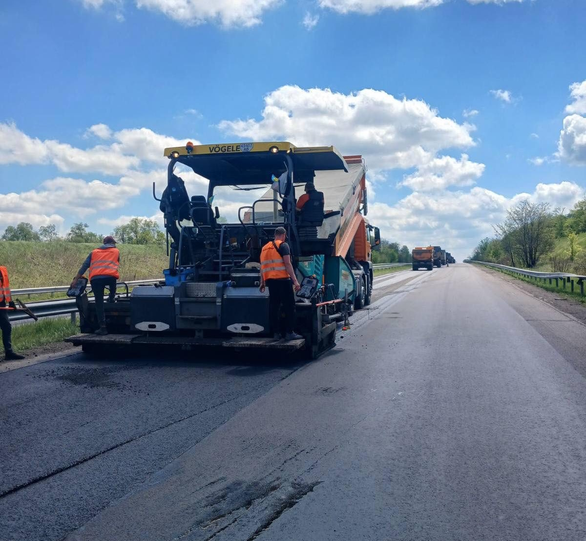Road paving machine laying fresh asphalt with workers in orange safety vests on a highway under a blue sky.