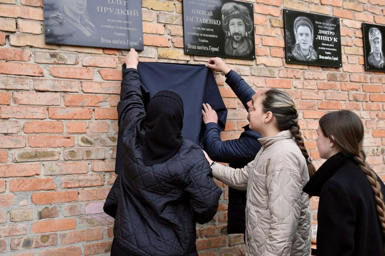 Three women unveil a memorial plaque on a brick wall by lifting a dark cloth.