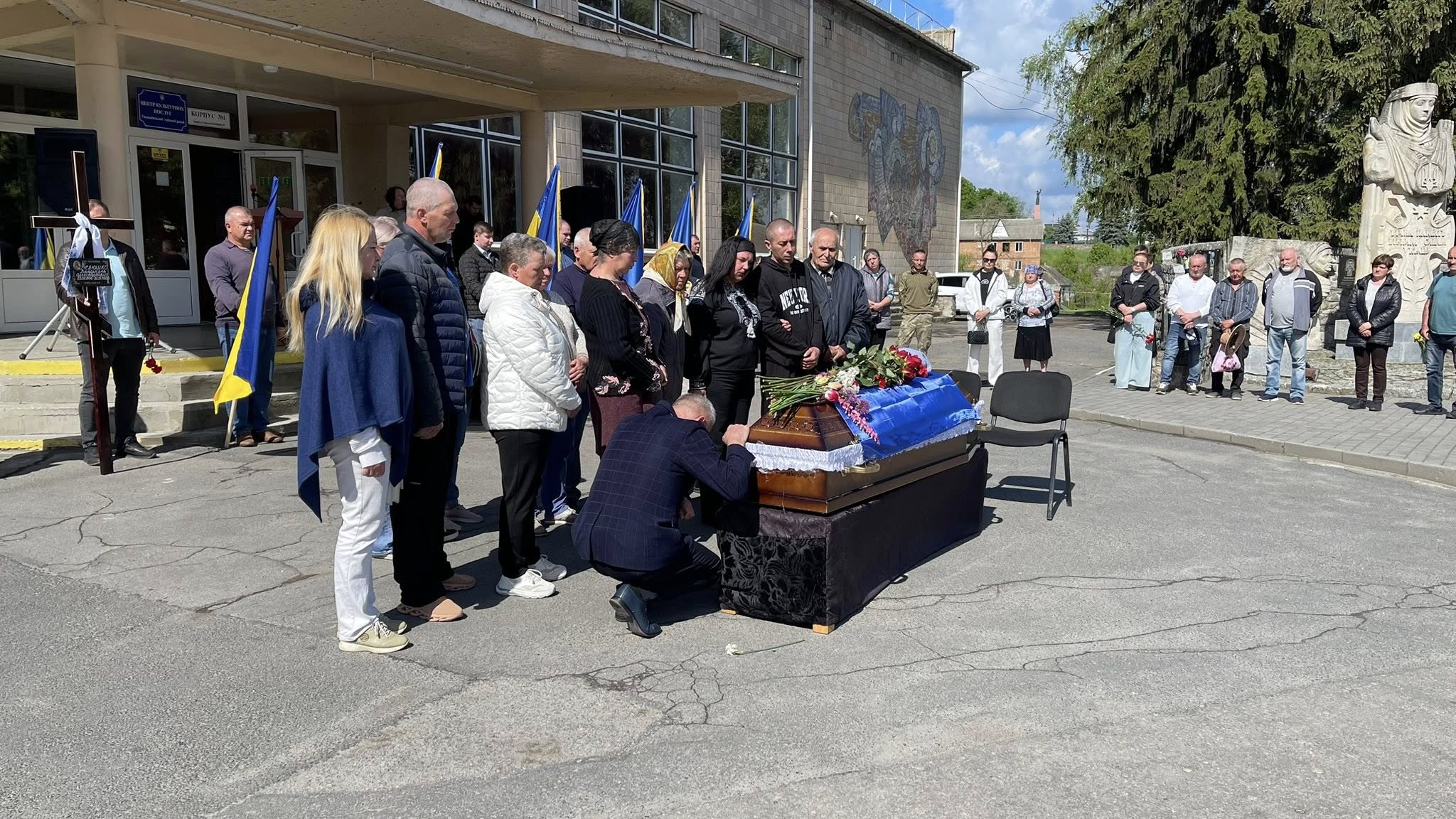 Mourners stand in a line beside a coffin draped in blue cloth with flowers, paying respects outside a building with Ukrainian flags nearby.
