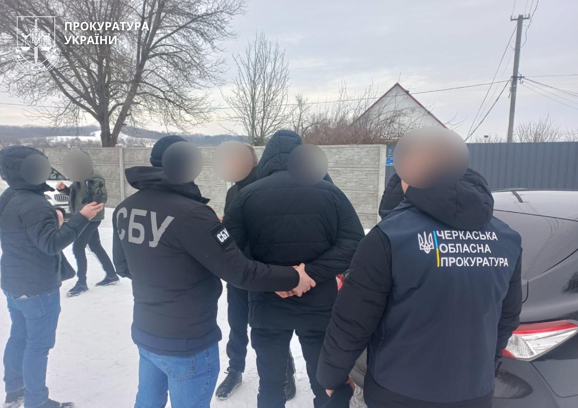 Group of people in winter jackets with СБУ insignia standing in a circle in a snowy outdoor area beside a car; a wall and utility poles in the background.