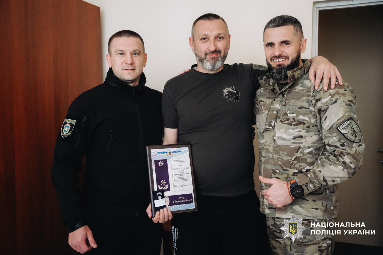 Three men pose indoors: a police officer in black uniform on the left, a civilian in the middle holding a framed award, and a soldier in camouflage on the right; Ukrainian police logo visible in the bottom right corner.