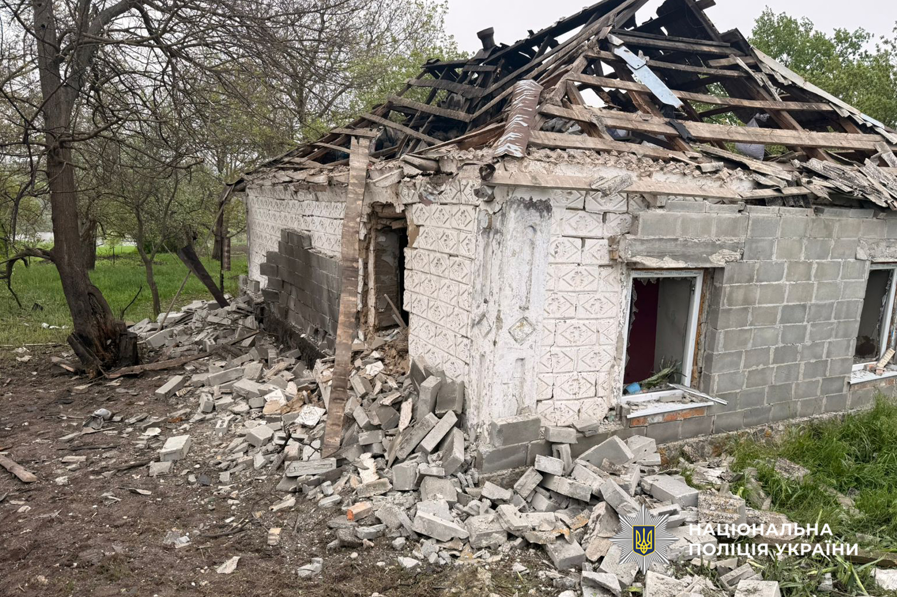 Collapsed house with a ruined roof and scattered concrete debris in a yard; trees nearby and a police watermark in the corner.