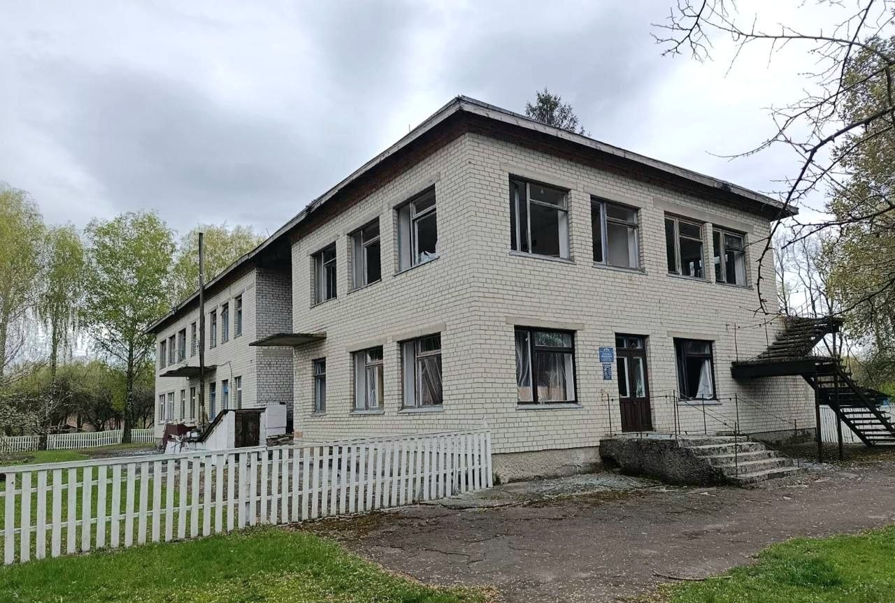 Two-story white brick building with external metal stairs, a white picket fence, and surrounding greenery on a cloudy day.