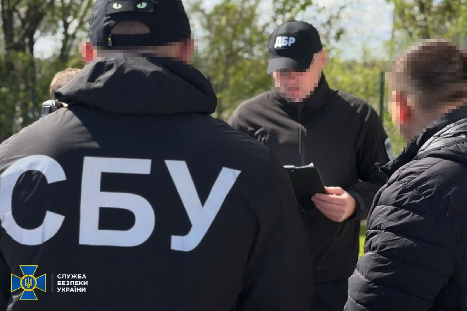 Group of people outdoors, two officers wearing black jackets with large 'СБУ' letters on the back, discussing with a man holding a notebook.