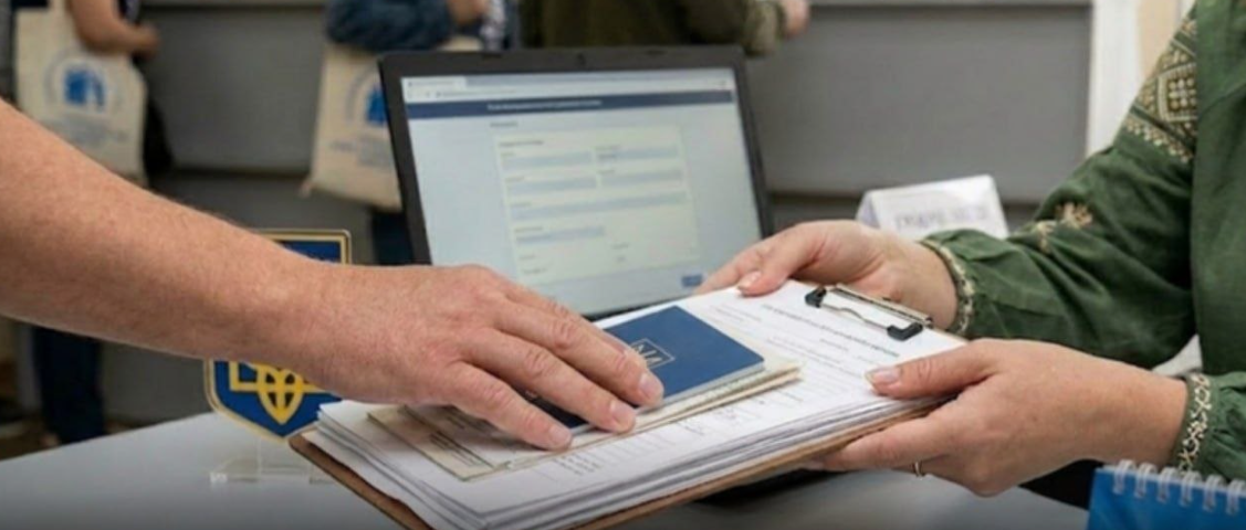 Person hands a blue passport and documents to another person across a desk, with a laptop in the background.