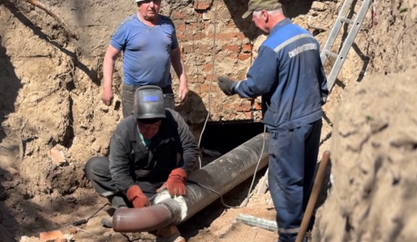 Three construction workers in a dirt trench repair a large metal pipe; one kneels and seals the joint while another watches, a ladder rests nearby.