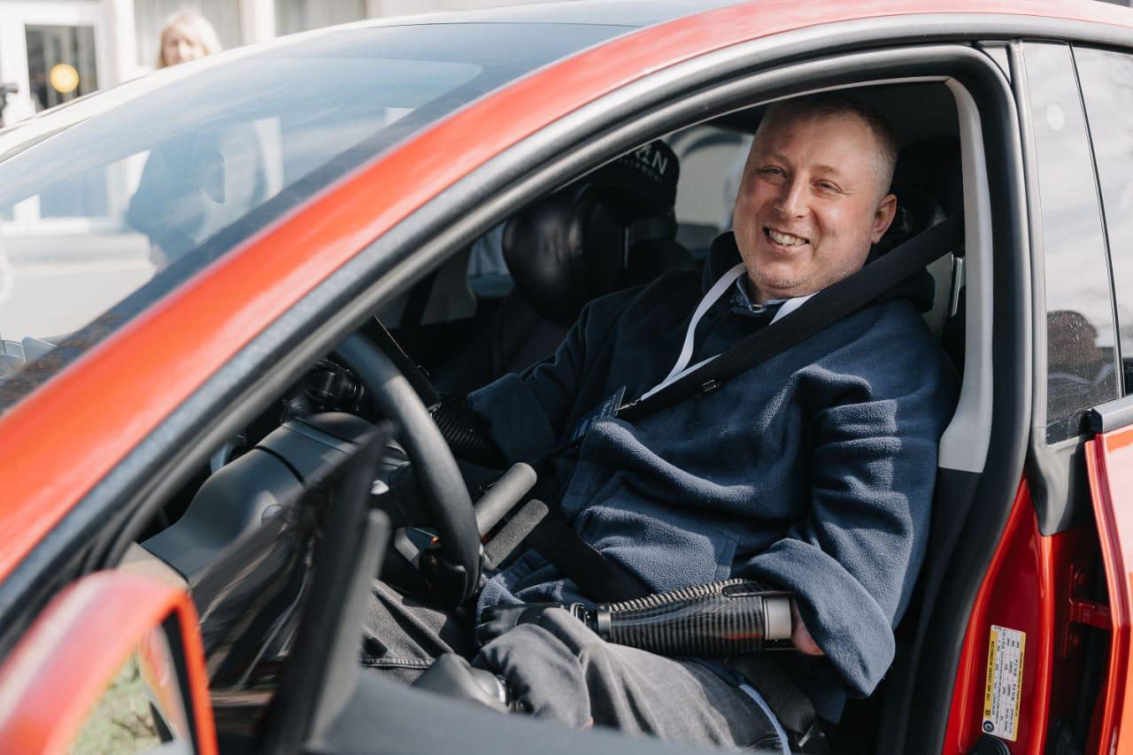 Smiling man sits in a red car, wearing a seatbelt and hoodie, ready to drive.