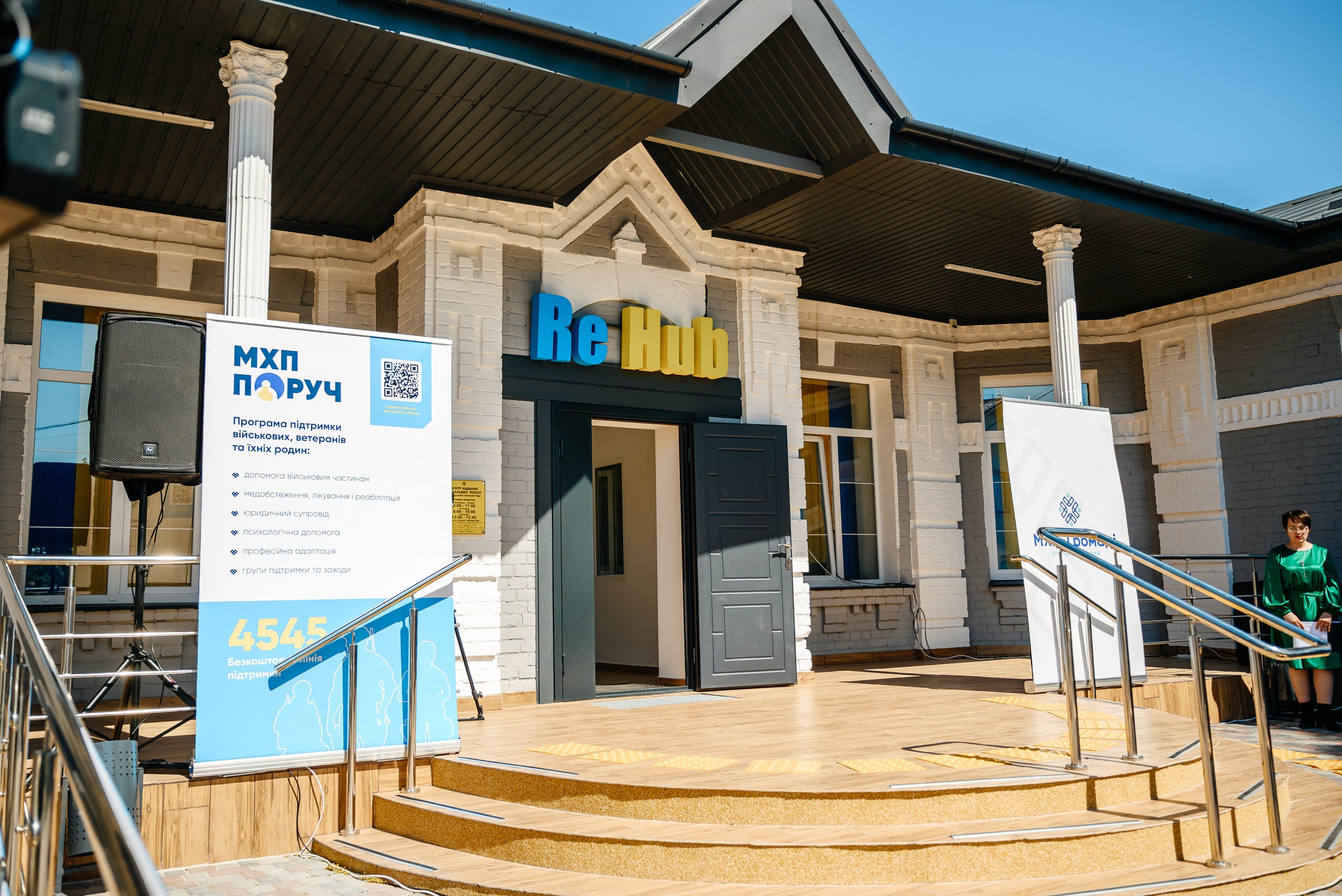 Exterior of a brick building with a blue-and-yellow 'Re Hub' sign above the entrance, banners set up outside, and a person in green standing to the right under a clear blue sky.